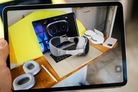Photo for Paris, France - Jun 6, 2023: An image on a iPad tablet showcases a wooden desk set up with a laptop displaying futuristic Vision Pro goggles, a striped VR headset resting atop the keyboard, paired ear - Royalty Free Image