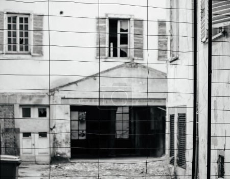 Téléchargez les photos : Image en noir et blanc d'une clôture métallique devant un bâtiment abandonné, illustrant le concept de désintégration urbaine - en image libre de droit
