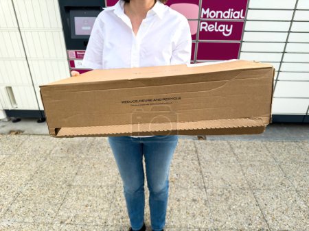Foto de París, Francia - 4 de octubre de 2023: Una mujer con una camisa blanca que sostiene una larga caja de cartón etiquetada REDUCE, REUSE, AND RECYCLE, de pie contra un telón de fondo de los casilleros Mondial Relay - Imagen libre de derechos