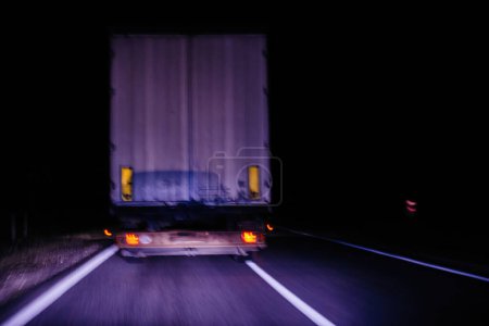 Téléchargez les photos : Capture floue du mouvement d'un camion circulant sur une autoroute la nuit, mettant en évidence les feux arrière et l'arrière du camion dans un fond sombre - en image libre de droit