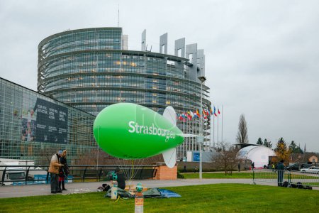 Photo for Strasbourg, France - Nov 24, 2014: Individuals set up a massive green dirigible balloon, marked with Strasbourg, before the Parliament building - Royalty Free Image