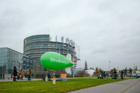Téléchargez les photos : Strasbourg, France - 24 nov. 2014 : Des gens s'affairent à préparer un vaste dirigeable écologique vert marqué Strasbourg devant le Parlement, présenté dans une perspective large et saisissante - en image libre de droit