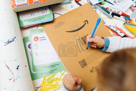 Foto de París, Francia - 25 de octubre de 2023: Desde arriba, un curioso niño dibuja los ojos en un logotipo de Amazon usando lápices de colores. En su mesa creativa, la escena sugiere un creciente interés en el diseño gráfico. - Imagen libre de derechos