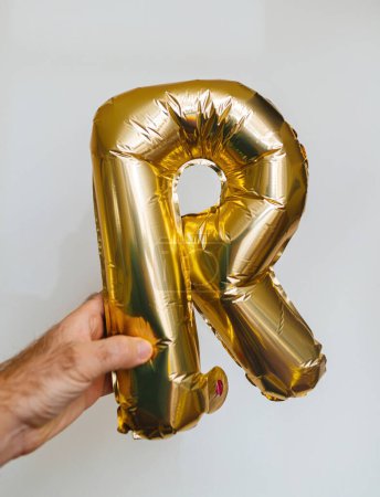 Téléchargez les photos : Une main masculine montre la lettre R, fabriquée à partir de ballons d'hélium dorés scintillants. Positionné sur un fond blanc pur, son parfait pour diverses célébrations - en image libre de droit
