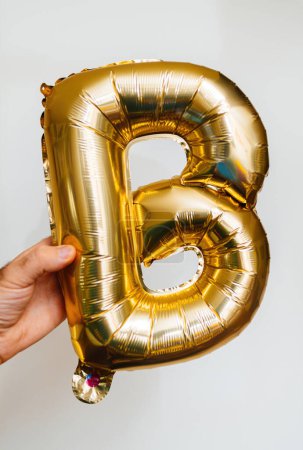 Foto de Una mano masculina sostiene la letra B, hecha a mano de brillantes globos de helio de oro, colocados sobre un fondo gris. - Imagen libre de derechos
