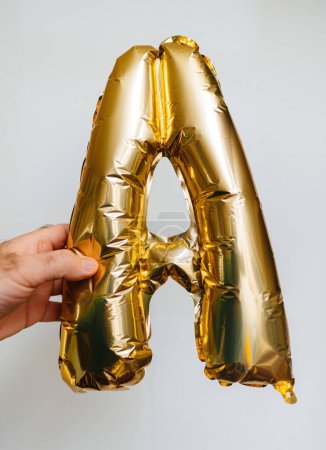 Foto de Una mano masculina sostiene la letra A, hecha de globos de helio de oro brillante, sobre un fondo blanco prístino - Imagen libre de derechos