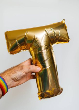 Foto de Una mano masculina sostiene la letra T, hecha de brillantes globos de helio dorado, sobre un fondo blanco - Imagen libre de derechos