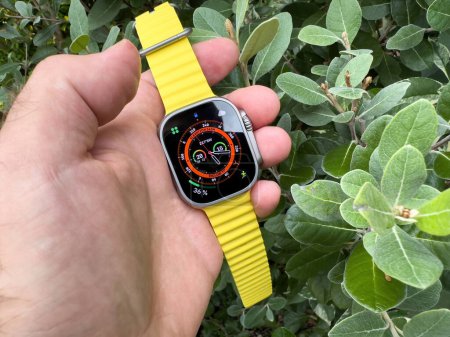 Photo for London, United Kingdom - Sep 23, 2023: A male hand holding the new Apple Watch Ultra 2 with a vibrant yellow rubber strap against a lush green background - Royalty Free Image
