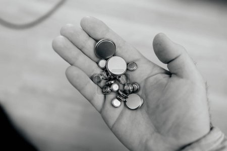 A black and white photograph captures a male hand clutching multiple button cell batteries, contrasting technology with timelessness