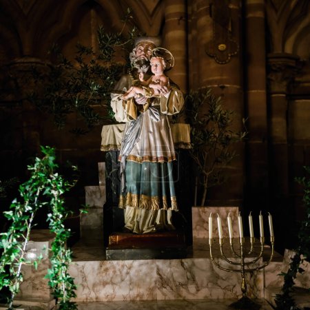 Téléchargez les photos : Représentation d'un saint tenant un enfant à Notre-Dame de Strasbourg, sur fond de chandelles et de feuillage pendant Noël - en image libre de droit