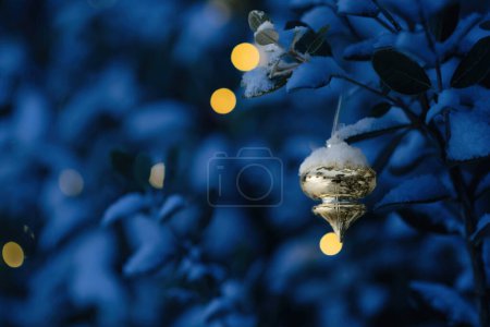 Foto de Un adorno festivo espolvoreado con nieve cuelga de una rama al atardecer, con luces amarillas brillando suavemente en el fondo - Imagen libre de derechos
