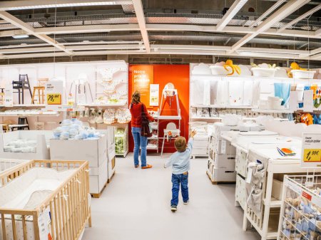 Téléchargez les photos : PAris, France - 31 août 2023 : Un tout-petit pointe avec impatience les accessoires et les objets de soins pour enfants dans un magasin IKEA, tandis que sa mère examine une chaise de bébé à proximité - en image libre de droit