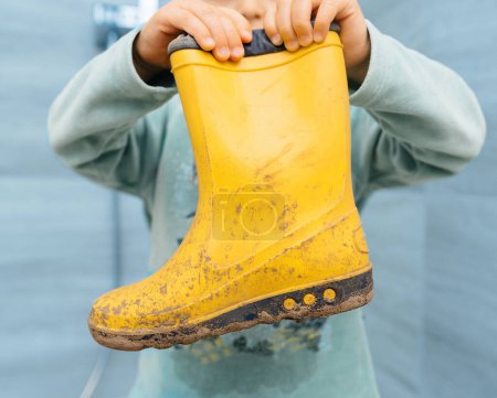 Foto de Un niño mostrando botas amarillas para caminar cubiertas de barro después de jugar al aire libre, simbolizando aventuras infantiles sin preocupaciones - Imagen libre de derechos