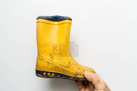 Foto de Una mano masculina sosteniendo botas de invierno otoñales amarillas al aire libre completamente cubiertas de barro, retratando el concepto de niñez sin preocupaciones y limpieza. - Imagen libre de derechos