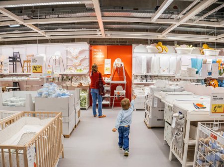 Téléchargez les photos : Paris, France - 31 août 2023 : Un bambin impatient pointe vers les accessoires et les objets de soins pour enfants dans IKEA, avec sa mère regardant une chaise bébé - en image libre de droit