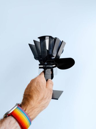 Foto de Mano masculina sosteniendo un ventilador de estufa negro e impotente, útil para mejorar la distribución de calor en una habitación, sobre un fondo azul - Imagen libre de derechos
