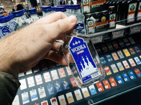 Foto de Frankfurt, Alemania - 10 de noviembre de 2023: Un disparo POV captura una mano masculina seleccionando cuidadosamente una botella de Vodka Maroska en un bullicioso supermercado alemán - Imagen libre de derechos