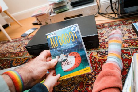 Foto de París, Francia - 16 de diciembre de 2023: Una mano masculina muestra el DVD Au Dodo - Ir a dormir junto a un pie de niño, con un reproductor de DVD Yamaha en el fondo. - Imagen libre de derechos