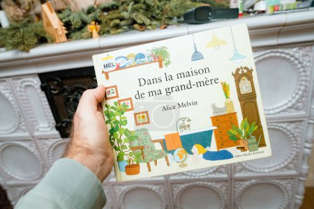 Foto de París, Francia - 16 de diciembre de 2023: Mano masculina presentando Dans la maison de ma grand-mere de Alice Melvin, un libro para niños, frente a una estufa rústica - Imagen libre de derechos