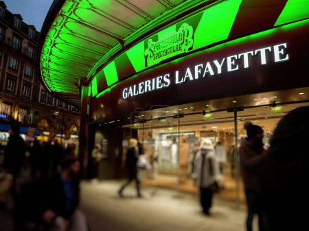 Téléchargez les photos : STRASBOURG, FRANCE - DEC 23, 2018 : L'emblématique signalisation des Galeries Lafayette brille au-dessus de l'entrée animée alors que les acheteurs flânent le long de la rue à la lumière du soir de Paris. - en image libre de droit
