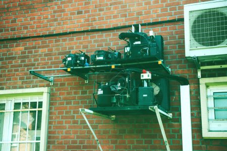 Téléchargez les photos : France - 10 juin 2016 : Plusieurs compresseurs hermétiques sont disposés à côté d'une thermopompe à air conditionné Daikin, face à une façade de maisons en brique, vue à faible angle - en image libre de droit