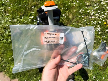 Téléchargez les photos : Francfort, Allemagne - 3 mai 2023 : Une personne tient un sac en plastique contenant des pièces pour l'assemblage de tondeuse à gazon, avec une nouvelle tondeuse à gazon et un champ herbeux parsemé de marguerites en arrière-plan. - en image libre de droit