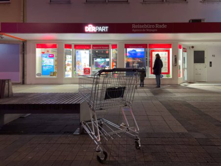 Foto de Kehl, Alemania - 1 de enero de 2024: Un carrito de compras solitario está abandonado en una tranquila acera por la noche, con la fachada iluminada de una agencia de viajes que proporciona un telón de fondo contrastante. - Imagen libre de derechos