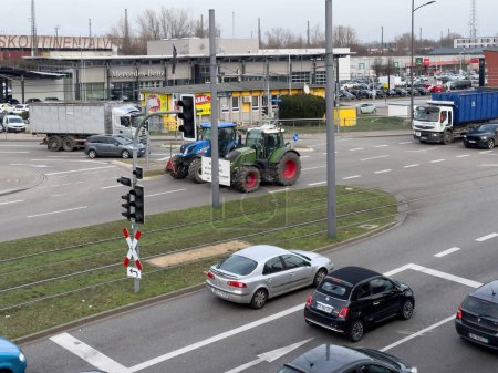 Foto de Kehl, Alemania - 9 de junio de 2024: Un tractor verde solitario se destaca entre el ajetreado flujo de automóviles y camiones en una carretera de la ciudad al atardecer. - Imagen libre de derechos