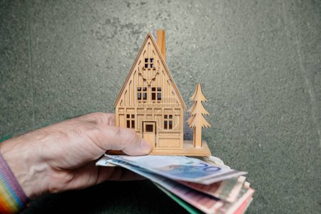 Photo for A man holds a small, wooden dream house against a stone background, accompanied by multiple euro banknotes, symbolizing aspirations for property ownership, real estate investment, or renting. - Royalty Free Image