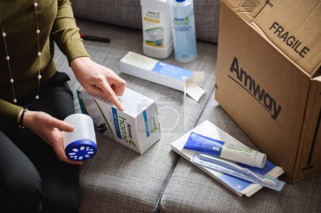 Foto de París, Francia - 20 de octubre de 2015: Mujer curiosa desempaqueta productos para el hogar de una caja Amway, organizando artículos como pasta de dientes y un dispensador de detergente en el sofá. - Imagen libre de derechos