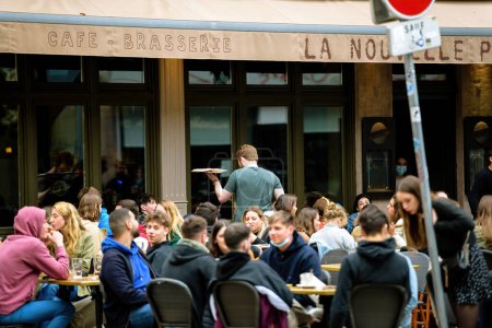 Téléchargez les photos : Strasbourg, France - 20 mai 2021 : Serveur avec tarte flambee food marchant entre les tables de Nouvelle poste terrasse pleine de personnes après l'épidémie de covidé-19 - en image libre de droit