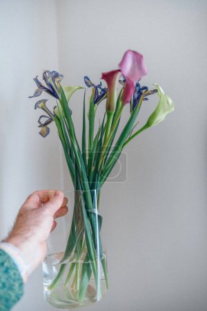 Foto de Una vibrante y colorida representación de una mano masculina delicadamente sosteniendo un jarrón lleno de lirios Cala y flores de iris, evocando la rica y animada esencia de este arreglo floral capturado en la película - Imagen libre de derechos