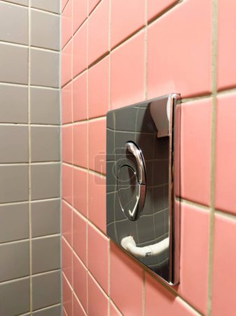 Foto de Un botón de descarga de inodoro brillante cromado instalado en la pared rosa con azulejos de la pared gris en contraste en el lado opuesto - Imagen libre de derechos