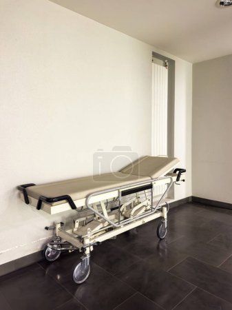 Téléchargez les photos : Image verticale d'un lit de transport d'hôpital vide sur le plancher de l'hôpital de la clinique avec des équipements de soins de santé en arrière-plan - en image libre de droit