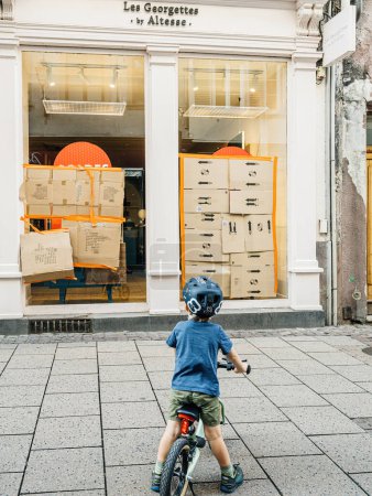 Photo for Strasbourg, France - Jul 2, 2023: Young child with helmet riding a bike and observing a storefront window with cardboard boxes. - Royalty Free Image