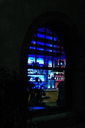Téléchargez les photos : Vue de nuit à travers une fenêtre voûtée dans l'intérieur bleu-éclairé d'un bar confortable. - en image libre de droit