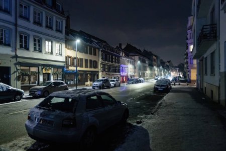 Foto de Estrasburgo, Francia - 23 de octubre de 2023: Coches y edificios cubiertos de nieve en una calle de la ciudad por la noche. - Imagen libre de derechos