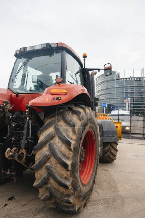 Téléchargez les photos : Strasbourg, France - 6 février 2024 : Vue latérale du tracteur rouge bloquant l'entrée du Parlement européen - protestation contre les importations de denrées alimentaires de moindre qualité et de nouveaux OGM. - en image libre de droit