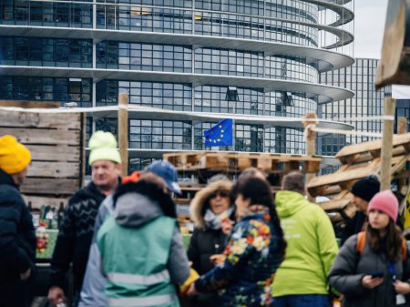 Téléchargez les photos : Strasbourg, France - 6 février 2024 : Des manifestants avec des tracteurs bloquent l'entrée au Parlement européen alors que des vagues de drapeau de l'UE défilent en arrière-plan, protestant contre les aliments importés avec des normes moins strictes et de nouveaux OGM - en image libre de droit