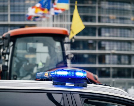 Foto de Estrasburgo, Francia - 6 de febrero de 2024: Baliza policial y visión desenfocada del tractor y el Parlamento Europeo mientras manifestantes con tractores bloquean la entrada, protestando contra alimentos importados con estándares más bajos - Imagen libre de derechos
