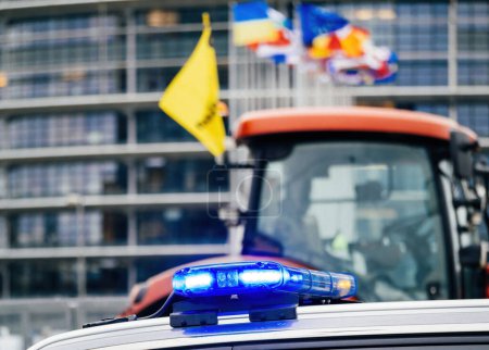 Photo for Strasbourg, France - Feb 6, 2024: Police beacon and defocused view of tractor and European Parliament as protesters block entrance, protesting imported food standards and new GMOs - Royalty Free Image