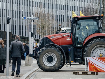 Téléchargez les photos : Strasbourg, France - 6 février 2024 : Un tracteur rouge entrave l'entrée de la rue au Parlement européen, protestant contre les importations de denrées alimentaires de moindre qualité et de nouveaux OGM - en image libre de droit