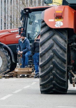 Téléchargez les photos : Strasbourg, France - 6 février 2024 : Des manifestants avec des tracteurs bloquent l'entrée du Parlement européen alors qu'un homme coupe des palettes pour le feu, protestant contre les aliments importés avec des normes plus basses et de nouveaux OGM - en image libre de droit