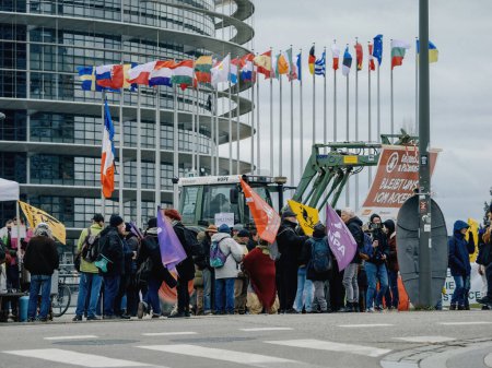 Foto de Estrasburgo, Francia - 6 de febrero de 2024: Manifestantes con tractores bloquean la entrada al Parlamento Europeo, se oponen a las normas alimentarias importadas y a los nuevos OMG, en medio de ondear banderas de los miembros de la UE - Imagen libre de derechos