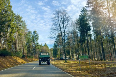 Téléchargez les photos : Breitenbrunnen, Allemagne - 27 déc. 2023 : De loin, un camion forestier vert Unimog conduit sur une route forestière, avec le panneau de la ville visible contre le ciel bleu clair. - en image libre de droit