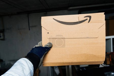 Photo for Bremen, Germany Dec 10, 2023: male hand presenting an Amazon Prime cardboard parcel in a large warehouse, showcasing the efficiency and convenience of online shopping and speedy delivery services. - Royalty Free Image