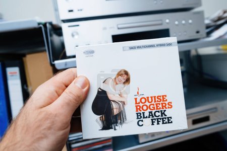 Téléchargez les photos : Paris, France - 28 déc. 2023 : main masculine tenant la nouvelle SACD de Louise Rogers intitulée Black Coffee a jazz Super Audio Compact Disk, avec un système audiophile haut de gamme en arrière-plan sur les étagères Vitsoe - en image libre de droit