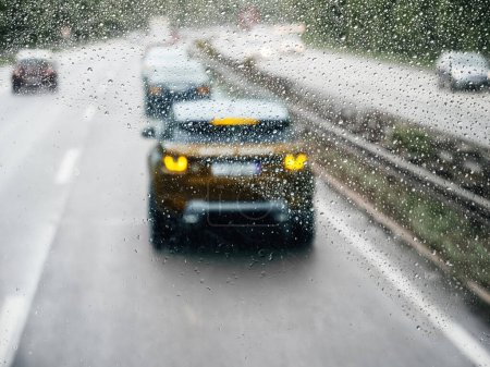 A rainy highway scene is seen from the perspective of a truck or bus at the front, with raindrops covering the windshield as vehicles drive along the road.