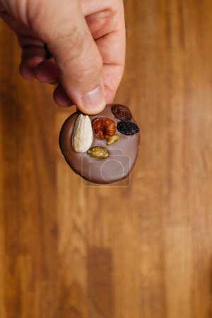 Eine männliche Hand hält zart einen mit Schokolade überzogenen Keks in der Hand, der mit verschiedenen Nüssen und getrockneten Trauben verziert ist und vor dem Hintergrund feiner Kunstfertigkeit ein köstliches Vergnügen darstellt..