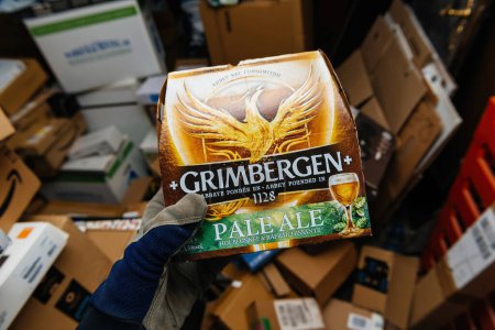Foto de Bremen, Alemania - 10 / 12 / 2023: En un gran garaje lleno de múltiples paquetes de cartón en el suelo, un hombre sostiene un paquete de cerveza Grimbergen Pale Ale, lo que sugiere un momento de relajación - Imagen libre de derechos